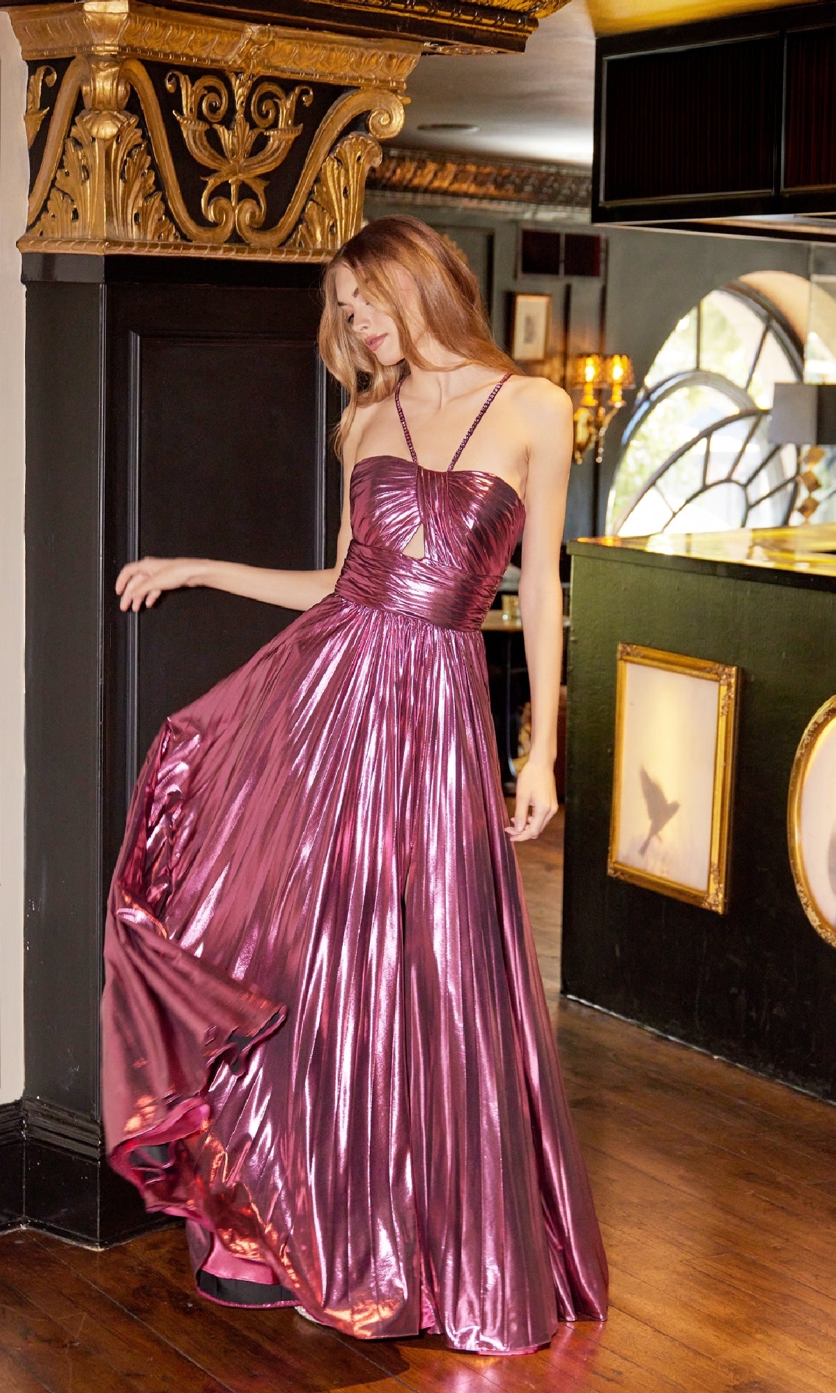 A woman indoors near a decorated column holds the side of her Long Metallic A-Line Prom Dress, Juliet JT2503A, featuring a shiny strappy-back design in metallic pink.